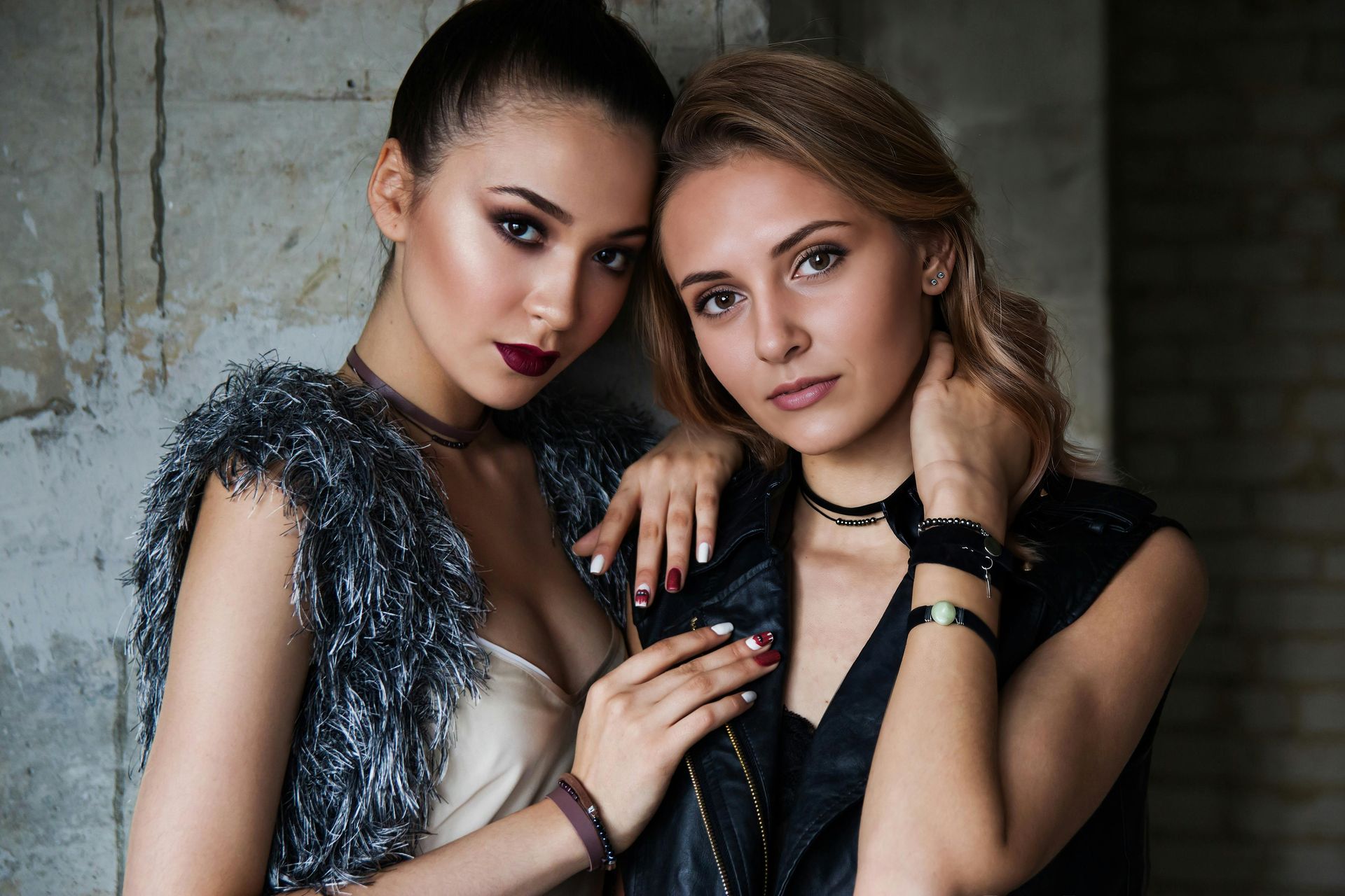 Two stylish women posing in edgy, high-fashion outfits against an industrial brick wall. One wears a black leather vest with statement accessories, while the other dons a faux fur vest, exuding confidence and modern glamour.
