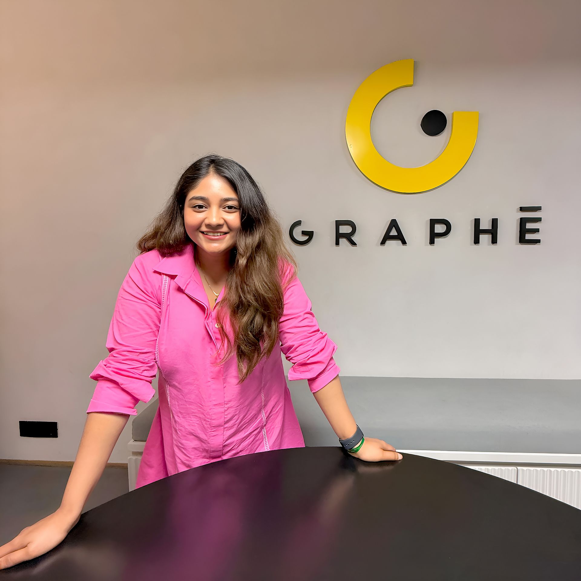 Yashashwi Malani smiling in front of the Graphē logo at her design studio, wearing a pink shirt and standing confidently.