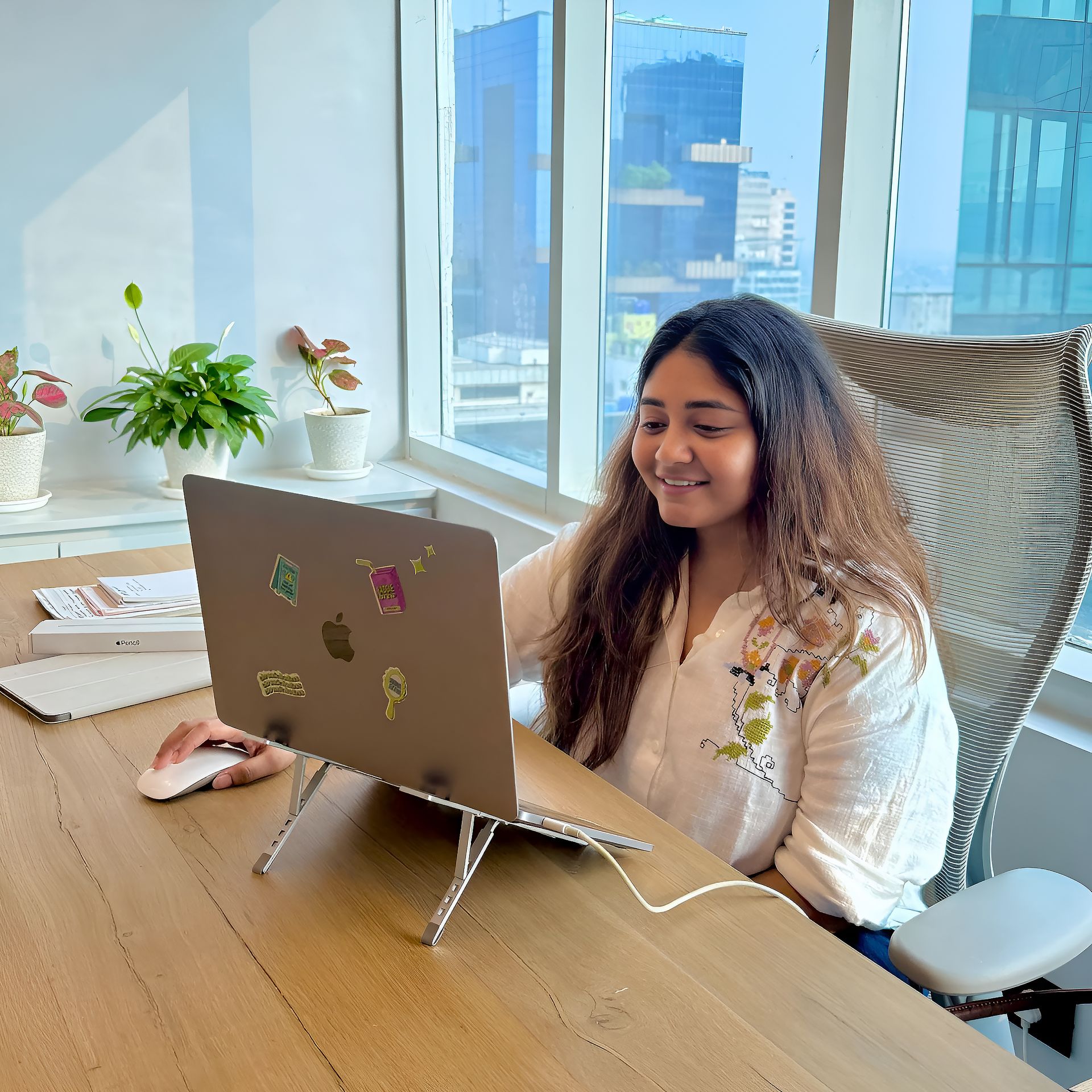 Yashashwi Malani working on a sticker-covered laptop at her office desk with indoor plants and a city view behind her, reflecting on saying yes to almost everything in her early career.