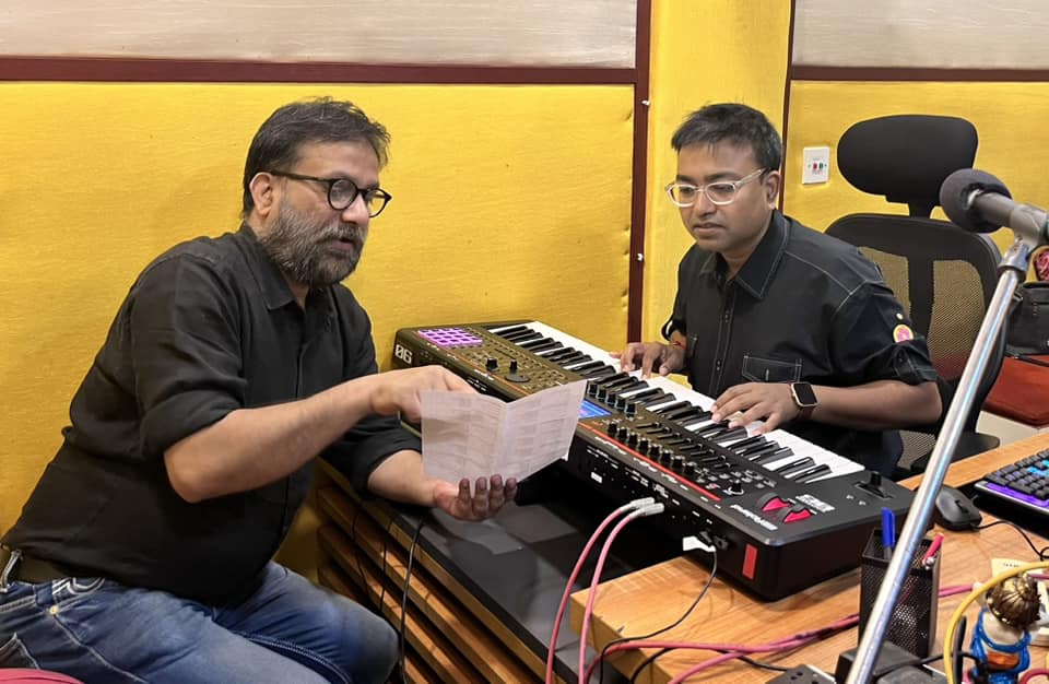 Drono Acharya and a colleague working together in a colorful recording studio, reviewing notes near a keyboard setup.